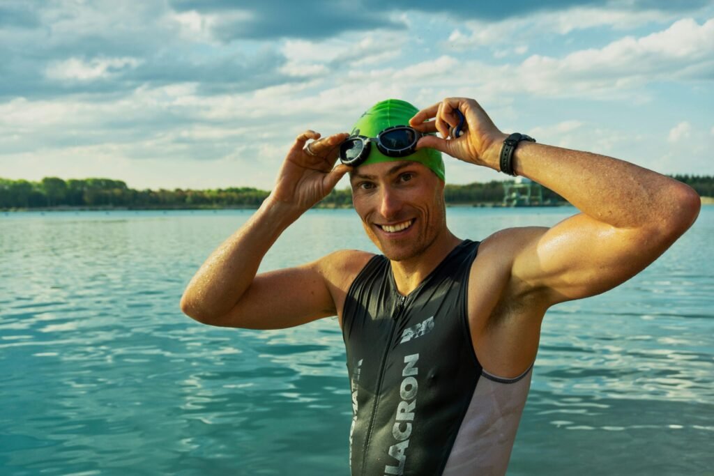A swimmer stands smiling in front of a large lake with a forested shoreline in the background. They wear a green swim cap and hold goggles above their eyes, dressed in a sleeveless wetsuit with visible “LACRON” and “NEW BALANCE” branding. The bright daylight and partly cloudy sky highlight the outdoor, open-water setting, capturing a moment of preparation or celebration in a swim event.